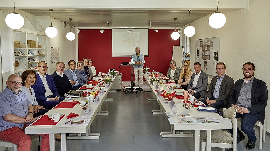 Foto: Menschen sitzen an Konferenztischen in einem Raum, im Hintergrund steht Herr Stritter an einem Podium vor einer Leinwand.