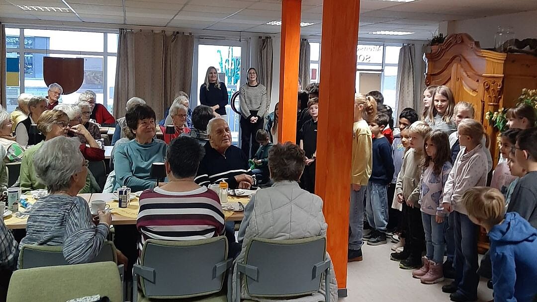 Foto: Senior*innen sitzen in einem Saal an langen Tischen, am Kopfende steht eine Gruppe Kinden