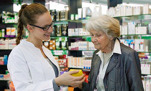 Eine Apothekerin zeigt einer älteren Frau in einer Apotheke eine Medikamentenschachtel