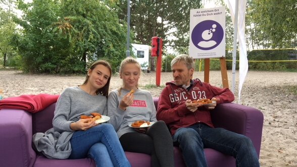 Foto: 3 Personen sitzen auf einem lila Sofa draußen auf einem Platz