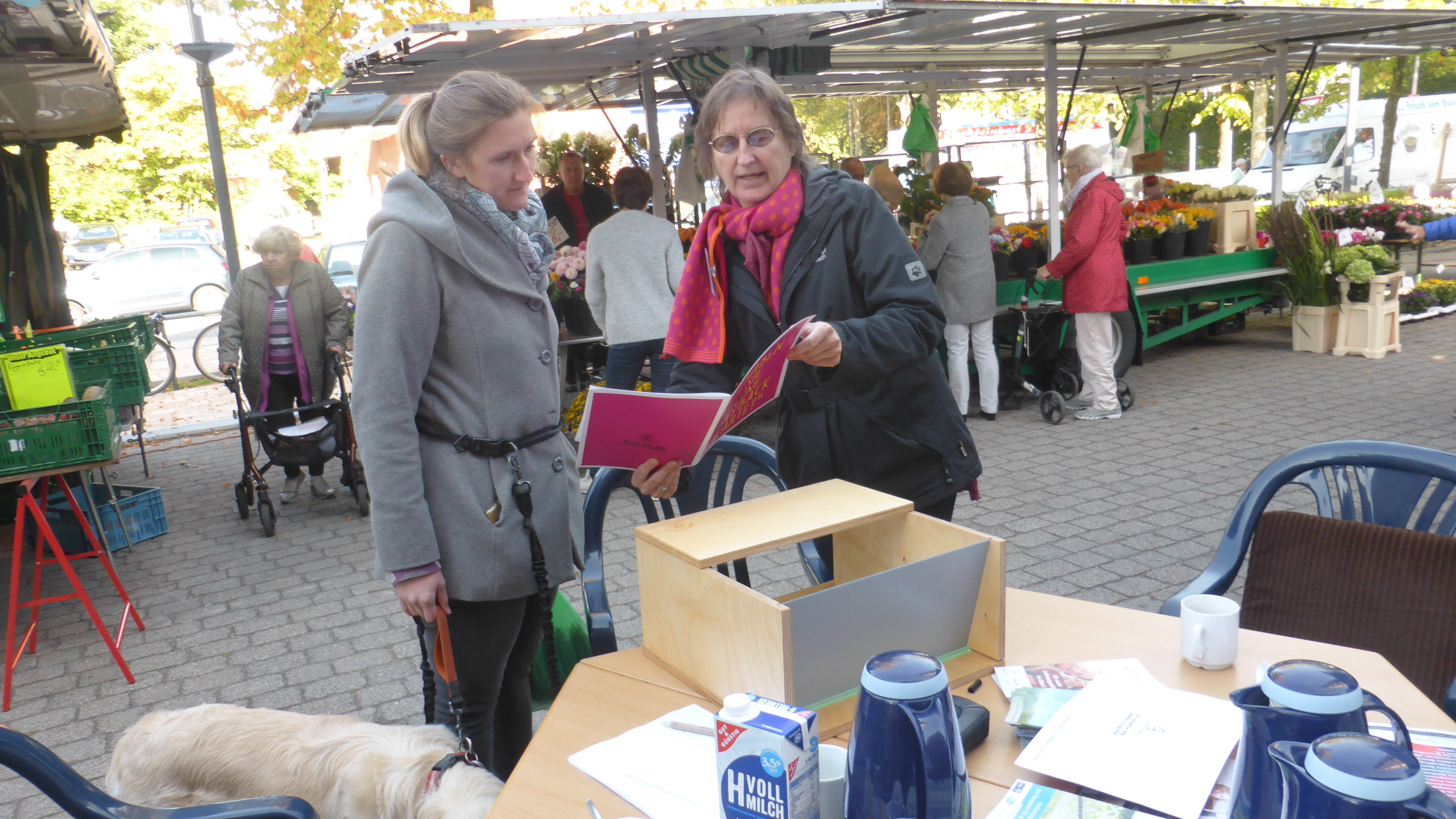 Eine Dame zeigt einer anderen Dame eine Publiaktion. Im Hintergrund ist ein Blumenstand.