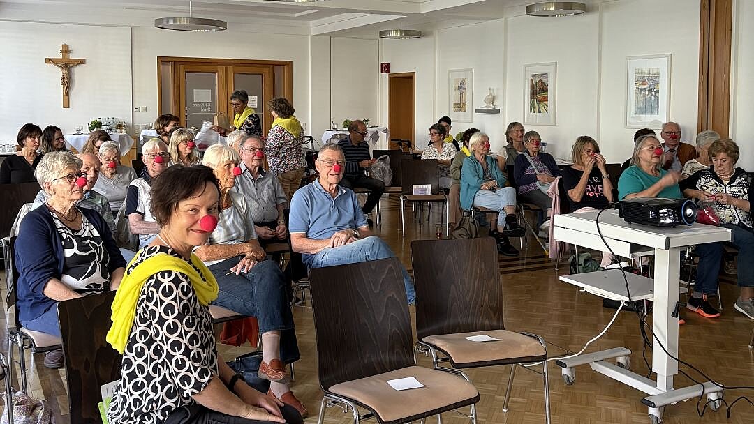 Foto: Senior*innen sitzen in einem Gemeindesaal und tragen rote Pappnasen