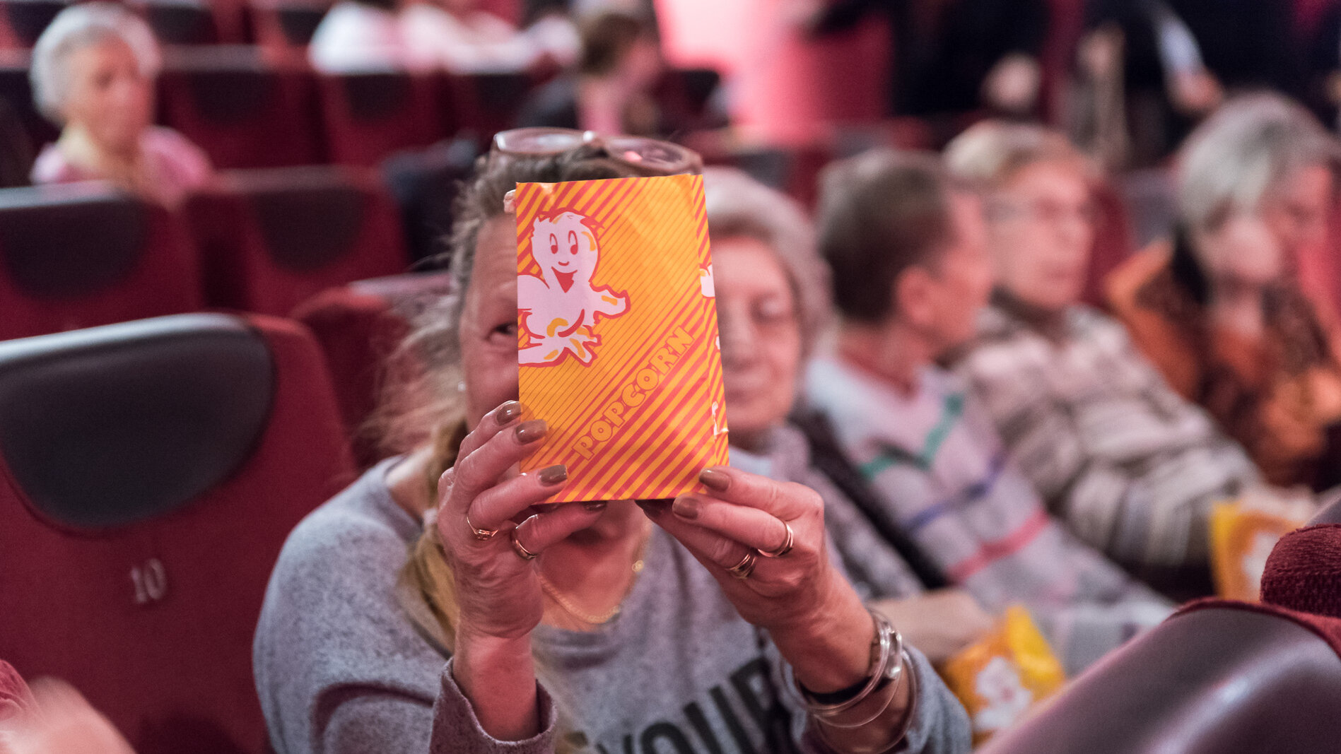 Auf diesem Foto sieht man ältere Menschen, die im Kono sitzen. Eine Frau im Vordergrund zeigt ihre Popkorntüte