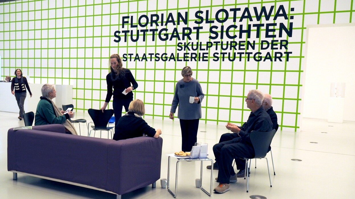 Foto: Sitzgruppe mit lila Sofa vor einer Wand mit Werbung für die Staatsgalerie Stuttgart