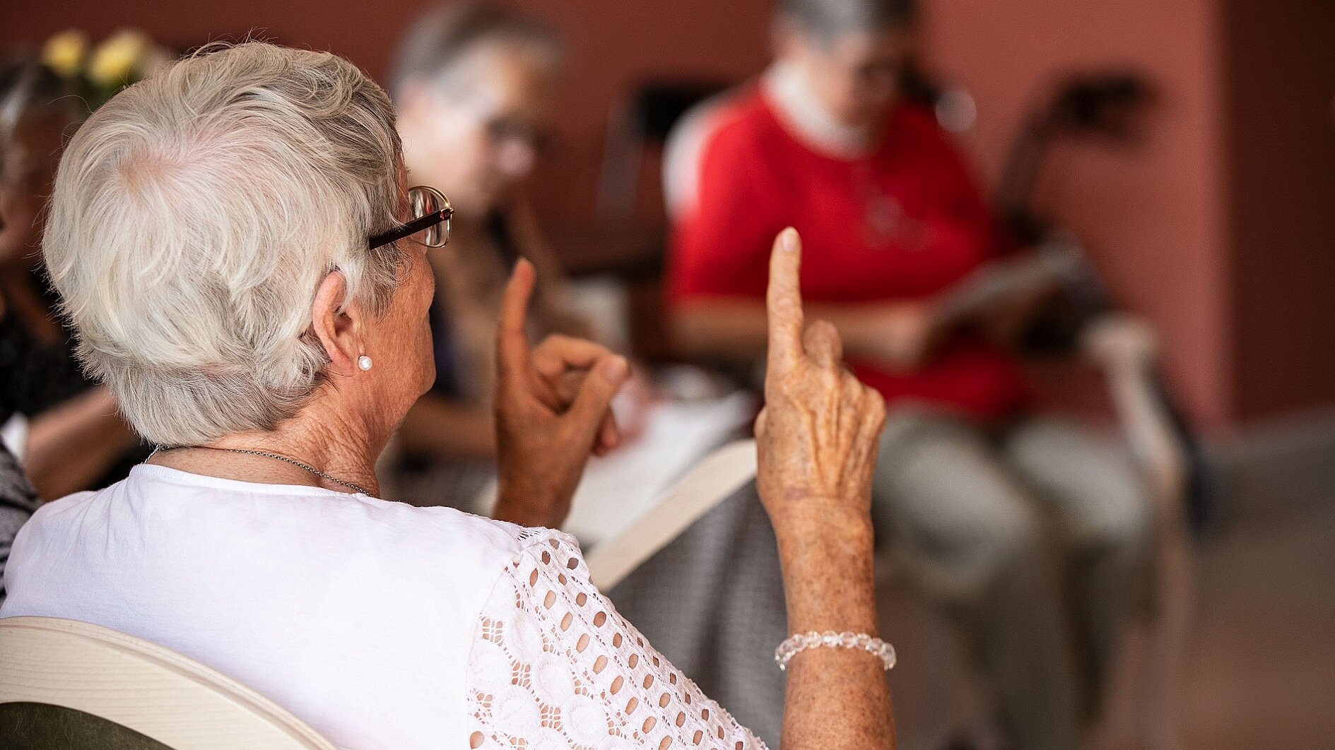 Foto: Ältere sitzende Frau von hinten, die ihre Zeigefinger in die Luft hält 