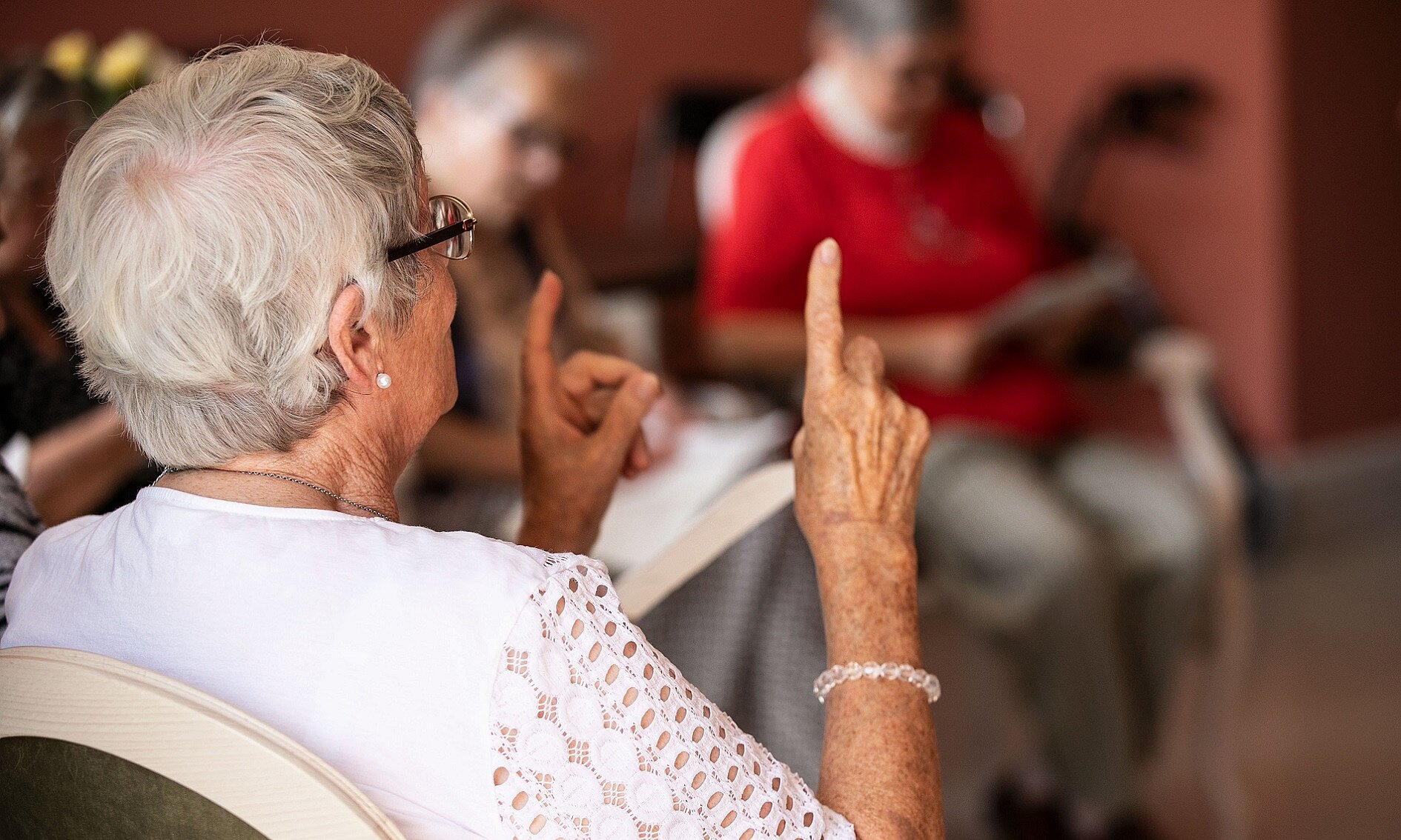 Foto: Ältere sitzende Frau von hinten, die ihre Zeigefinger in die Luft hält 