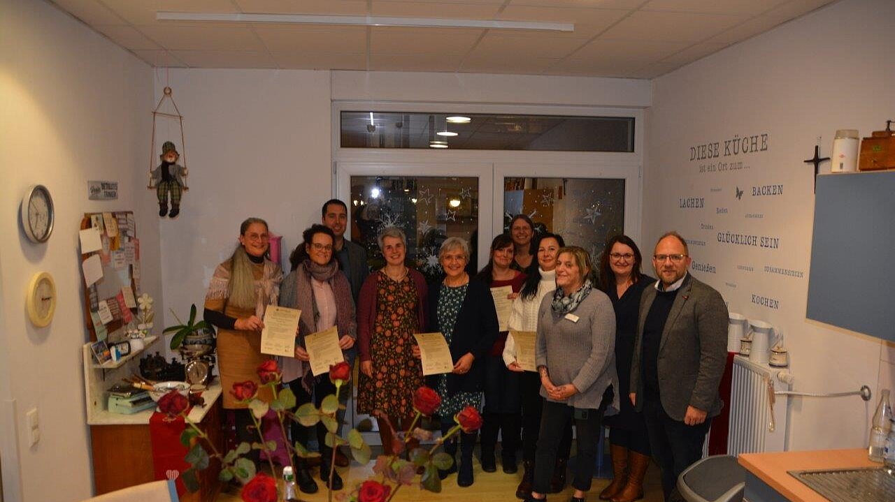 Foto: Gruppenbild von 11 Personen in einer Küche, die Bescheinigungen in der Hand halten. Im Vordergrund ein Strauß roter Rosen