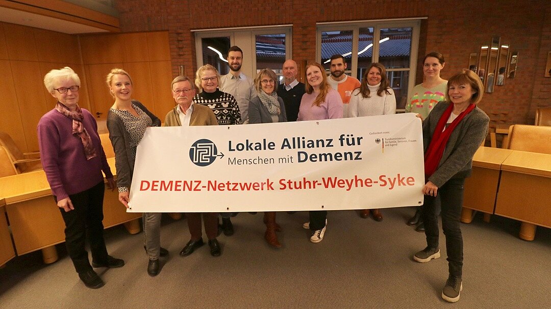Gemeinde Weyhe: Netzwerktreffen im Januar 2024 12 Personen stehen in einem Raum, 2 im Vordergrund halten ein großes Banner "DEMENZ-Netzwerk Stuhr-Weyhe-Syke"