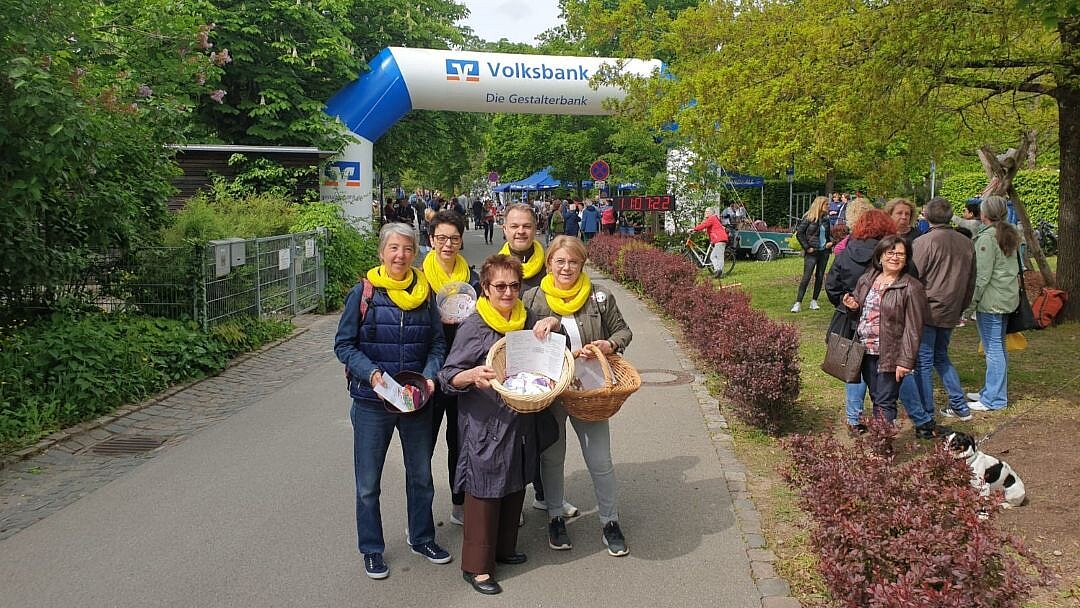 Foto: 5 Senior*innen mit gelben Schals auf einem Weg in einem Par, im Hintergrund Pavillons und viele weitere Personen
