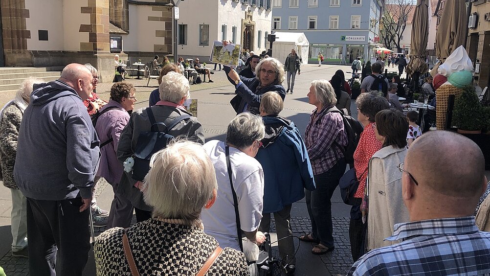 Foto: Eine Gruppe Senior:innen stehen auf einem Marktplatz um einen Mann herum, der ein großes Foto hochhält.