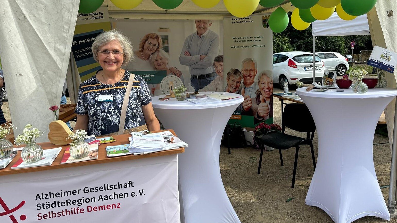 Foto: Seniorin steht hinter einem Infotresen mit Broschüren vor einem Pavillon mit weißen Stehtischen und grünen und gelben Luftballons unter dem Dach auf einem Parkplatz.