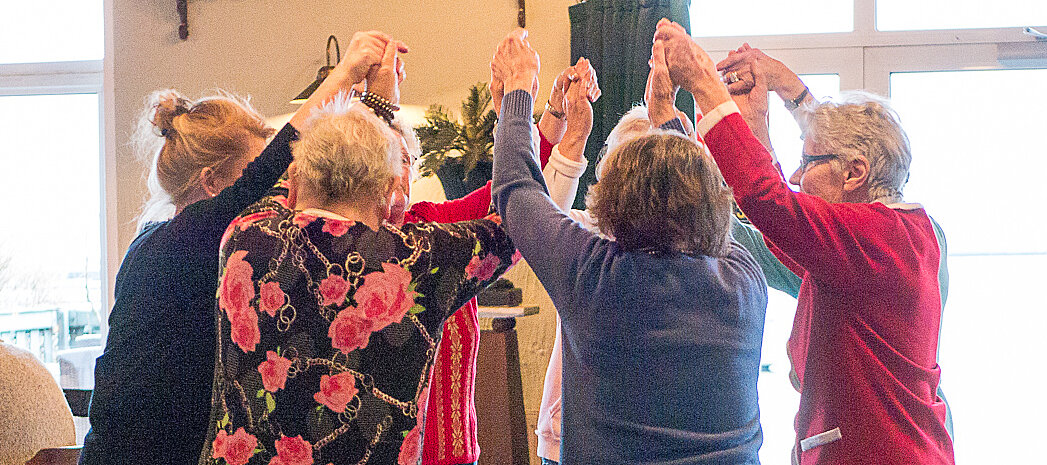 Foto: Sieben Personen stehen im Kreis und strecken die Arme hoch. Dabei halten sie sich an den Händen fest.