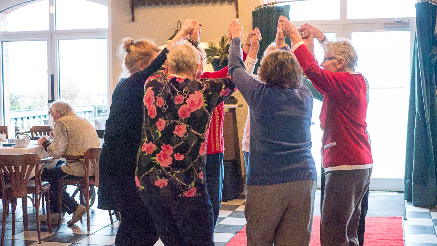 Foto: Sieben Personen stehen im Kreis und strecken die Arme hoch. Dabei halten sie sich an den Händen fest.