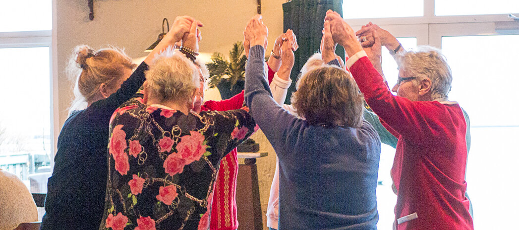 Foto: Sieben Personen stehen im Kreis und strecken die Arme hoch. Dabei halten sie sich an den Händen fest.