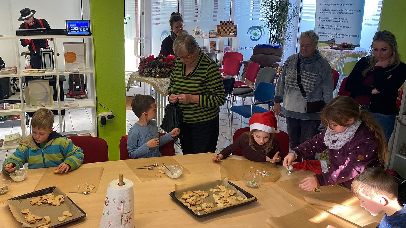 Foto: Kinder sitzen an einem großen Holztisch und backen Weihnachtsplätzchen, eine Erwachsene steht daneben und hilft. Im Hintergrund schauen drei Frauen zu.