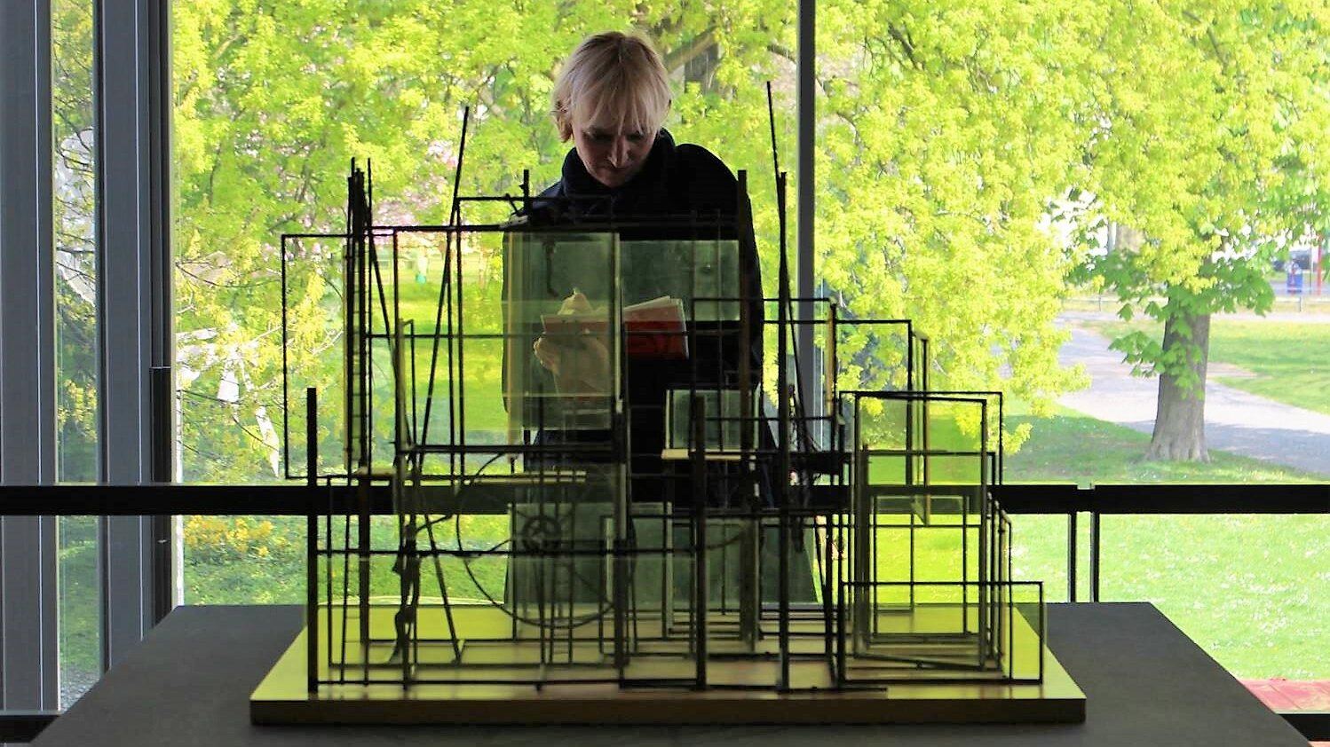 Im Vordergrund dieses Fotos sieht man ein abstraktes Kunstobjekt aus Glas, im Hintergrund steht eine Frau, die sich Notizen macht