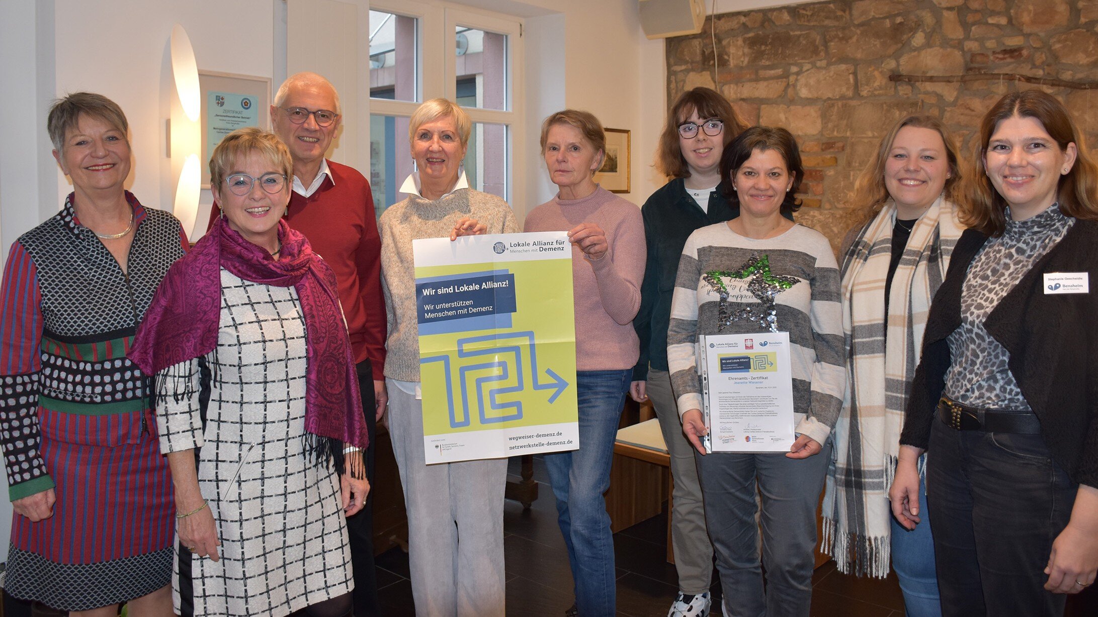 Foto: 9 Personen stehen vor einem Fenster, 2 halten ein Plakat der Lokalen Allianzen, eine hält das überreichte Zertifikat.