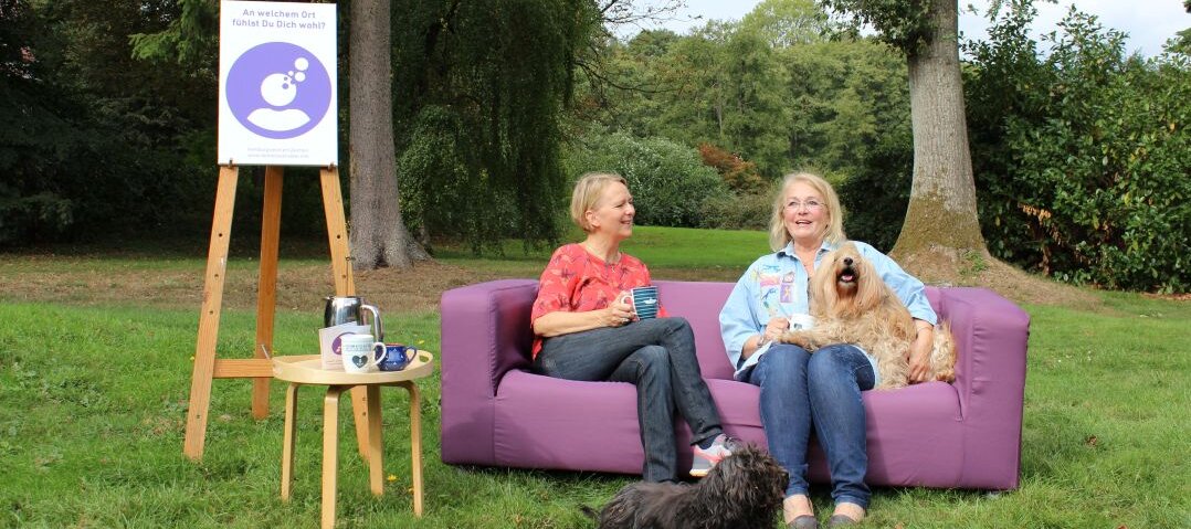 Auf diesem Foto sehen Sie ein lila Sofa, das draußen auf einer Wiese steht. Auf dem sofa sitzen zwei Frauen sowie zwei Hunde