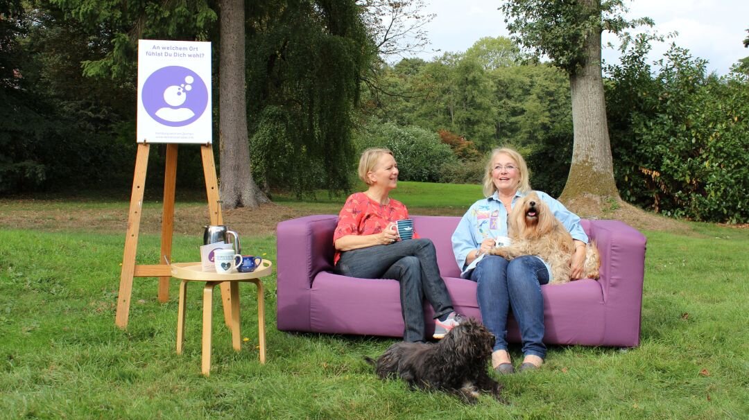 Auf diesem Foto sehen Sie ein lila Sofa, das draußen auf einer Wiese steht. Auf dem sofa sitzen zwei Frauen sowie zwei Hunde