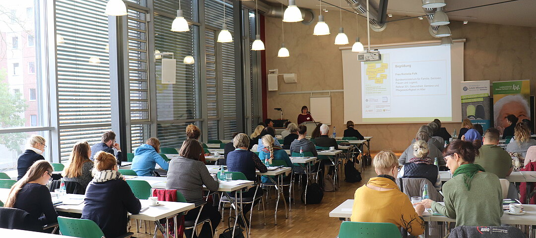 Teilnehmende sitzen in Tischreihen und schauen sich eine Präsentation auf einer Leinwand an