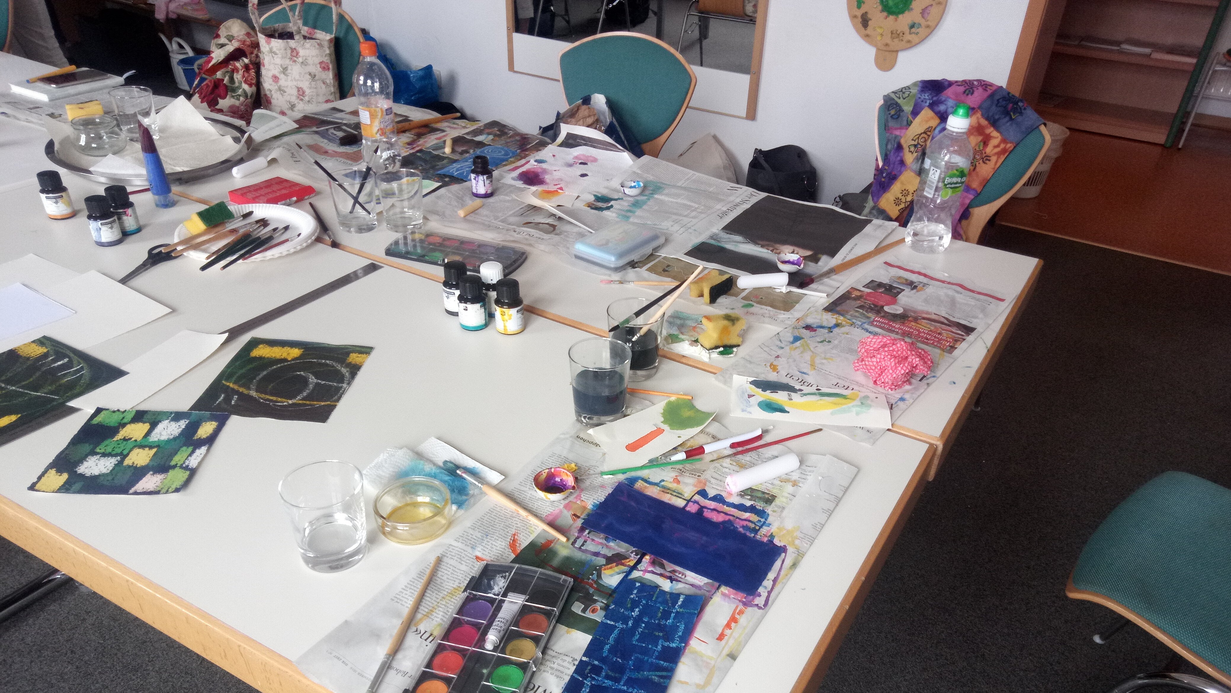 Foto: Großer Tisch, auf dem verschiedene Malutensilien sowie gerade gemalte Bilder liegen