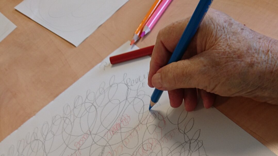 Auf dem Foto sieht man die Hände einer älteren Person, die ein abstraktes Bild zeichnet