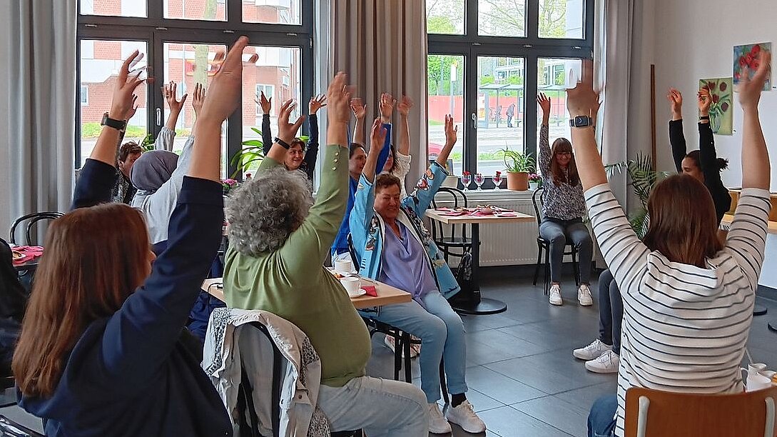 Stuhlgymnastik bei der Informationsveranstaltung Foto: Senioren sitzen in einem Stuhlkreis in einem Raum und heben die Arme in die Höhe.