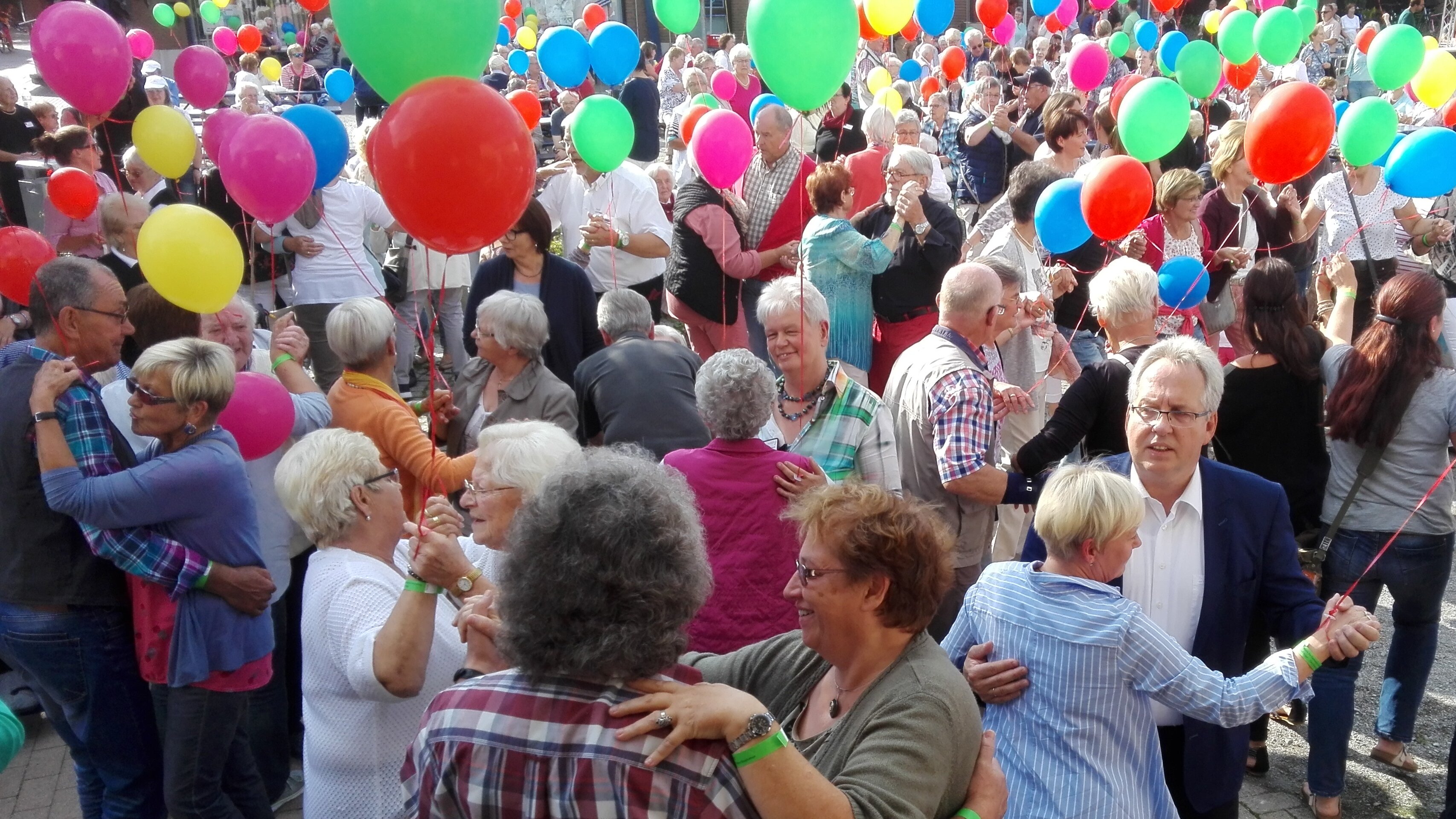 Auf diesem Bild sehen Sie einen Platz, auf dem viele ältere Menschen miteinander tanzen. Viele von ihnen haben einen Luftballon in der Hand.