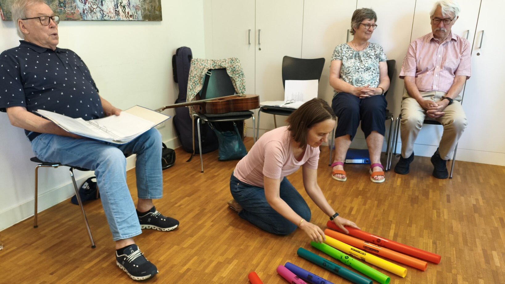 Foto: 3 Senior*innen sitzen auf Stühlen um eine junge Frau, die am Fußboden knieend Klangröhren in Regenbogenfarben spielt
