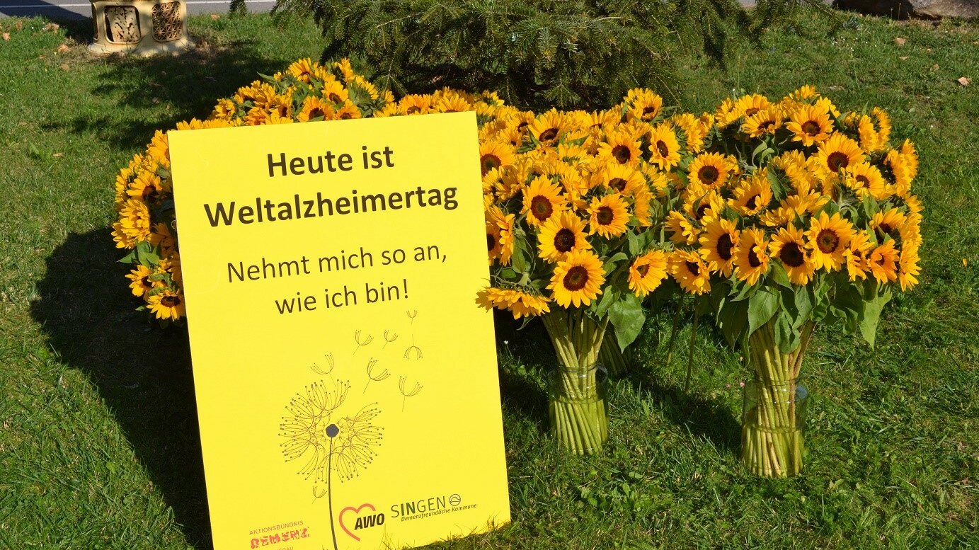 Vasen mit Sonnenblumen. Davor steht ein gelbes Plakat mit der Aufschrift "Heute ist Weltalzheimertag!"