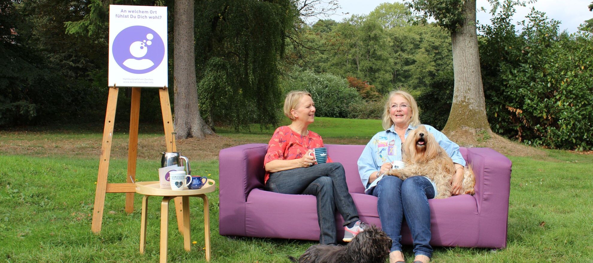 Auf diesem Foto sehen Sie ein lila Sofa, das draußen auf einer Wiese steht. Auf dem sofa sitzen zwei Frauen sowie zwei Hunde