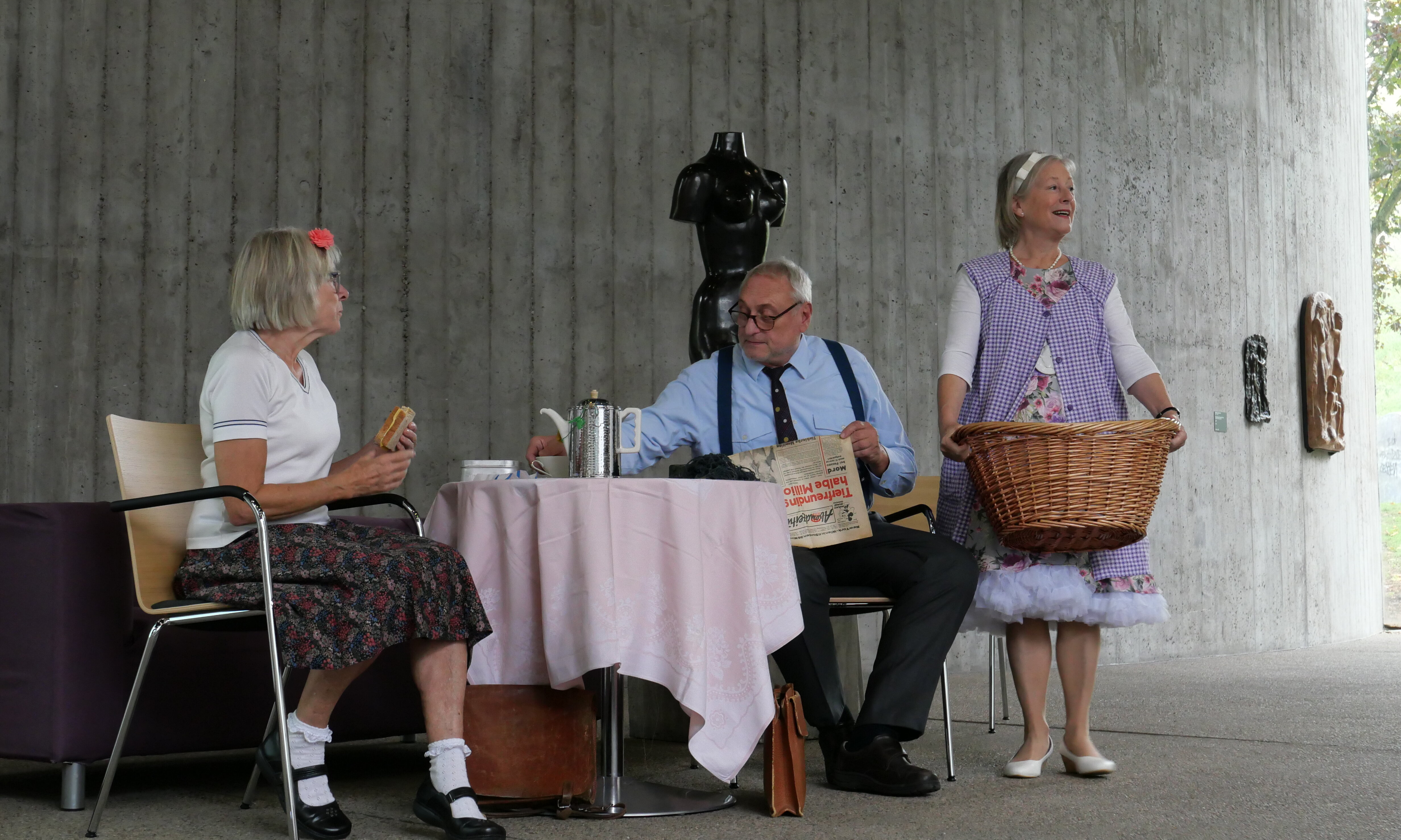 Theaterstück auf der Bühne: Zwei Personen sitzen am Tisch, eine Person hält einen Wäschekorb in der Hand