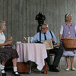 Theaterstück auf der Bühne: Zwei Personen sitzen am Tisch, eine Person hält einen Wäschekorb in der Hand