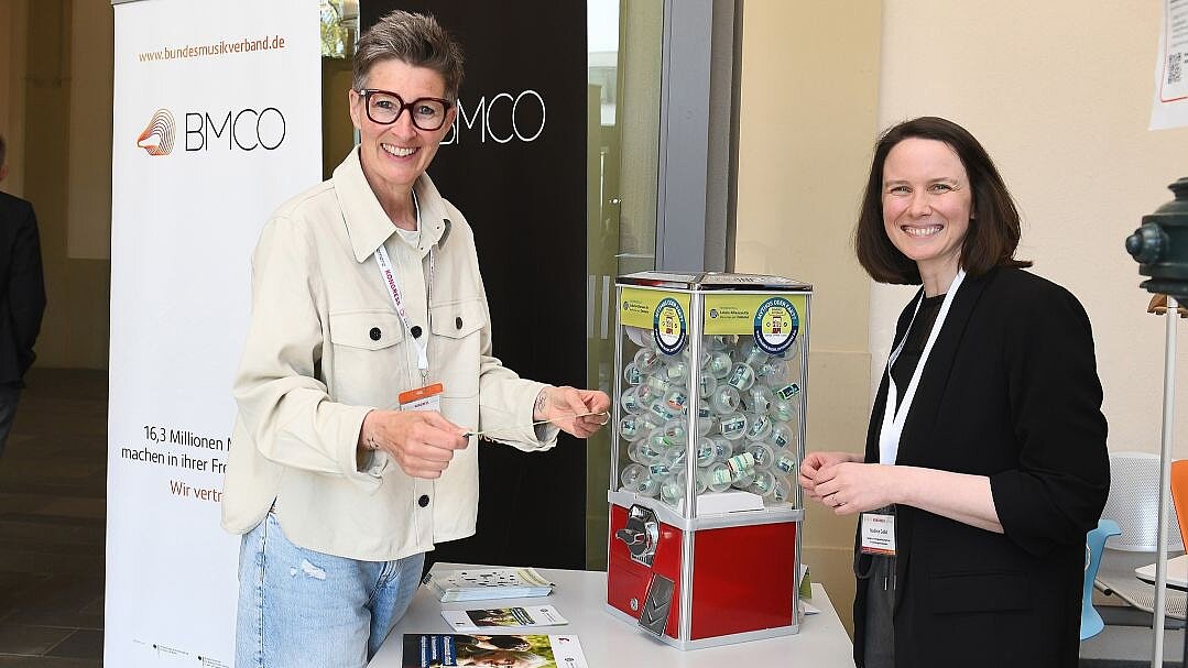 Foto: Zwei Frauen an einem Messestand, in der Mitte ein Kaugummiautomat und Broschüren auf einem Tisch.