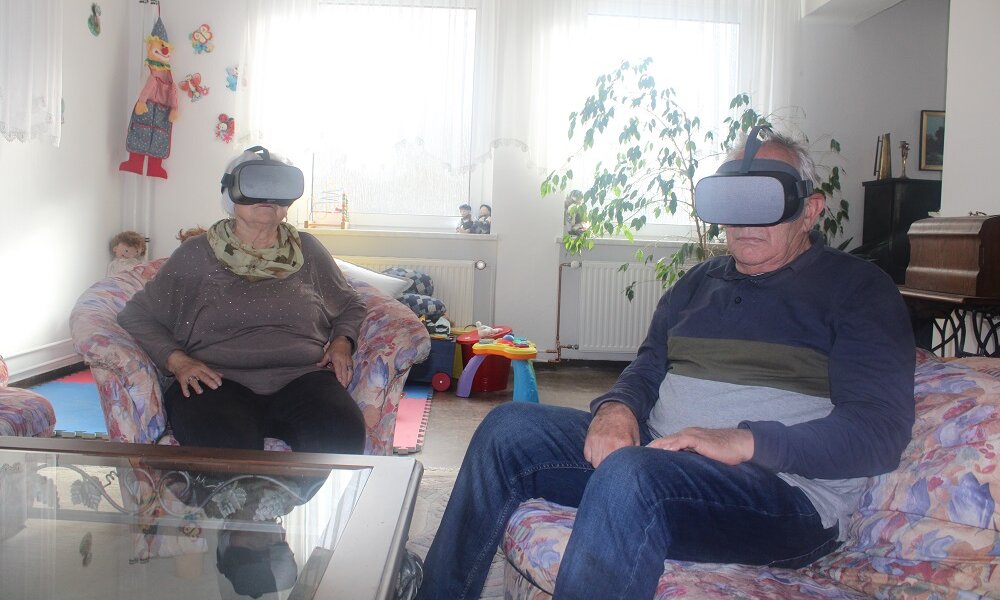 Eine ältere Frau und ein älterer Mann sitzen in einer Sitzgruppe und tragen Virtual-Reality-Brillen 