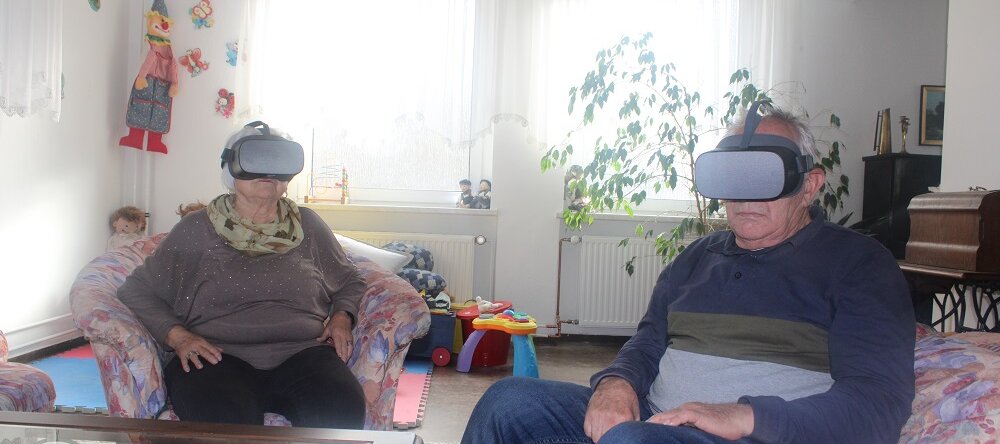 Eine ältere Frau und ein älterer Mann sitzen in einer Sitzgruppe und tragen Virtual-Reality-Brillen 