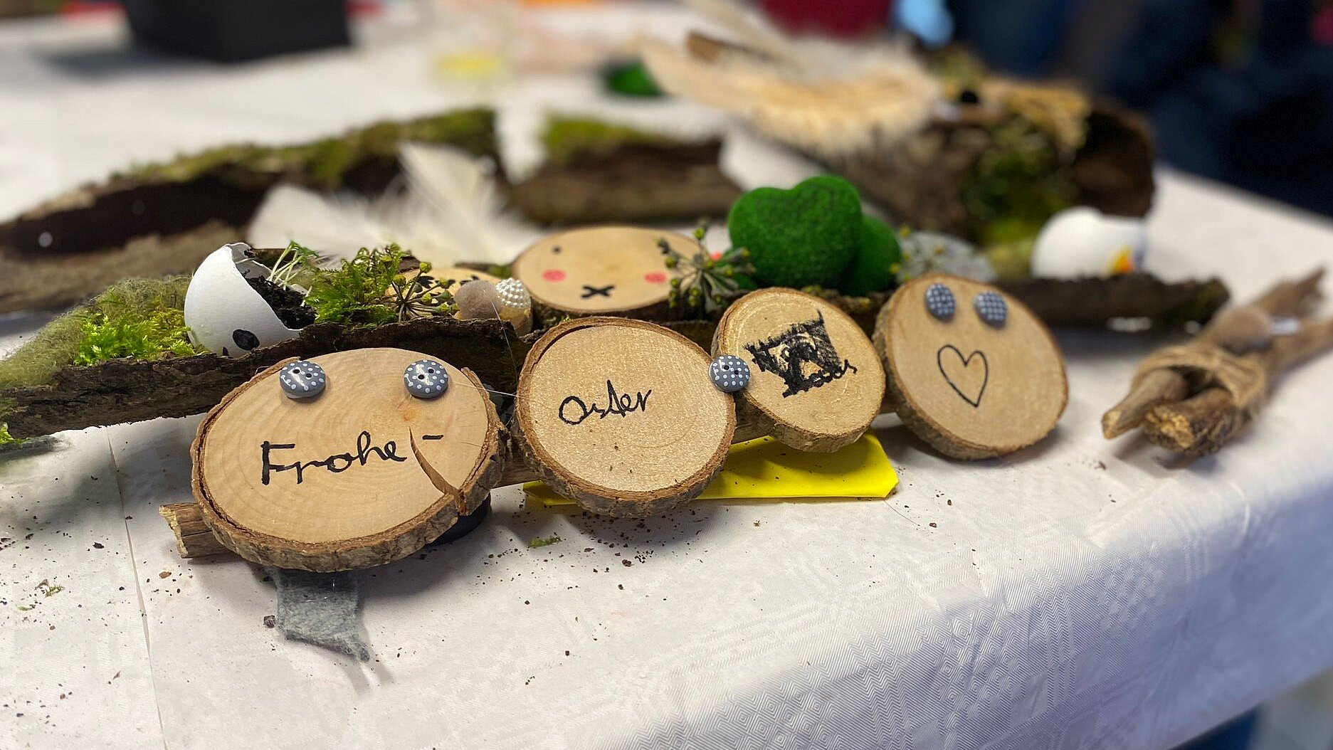 Foto: Auf einem Tisch mit weißer Tischdecke liegen Holzscheiben, mit Knöpfen und Moos dekoriert und mit schwarzem Stift mit "Frohe Ostern" und einem Herz beschriftet