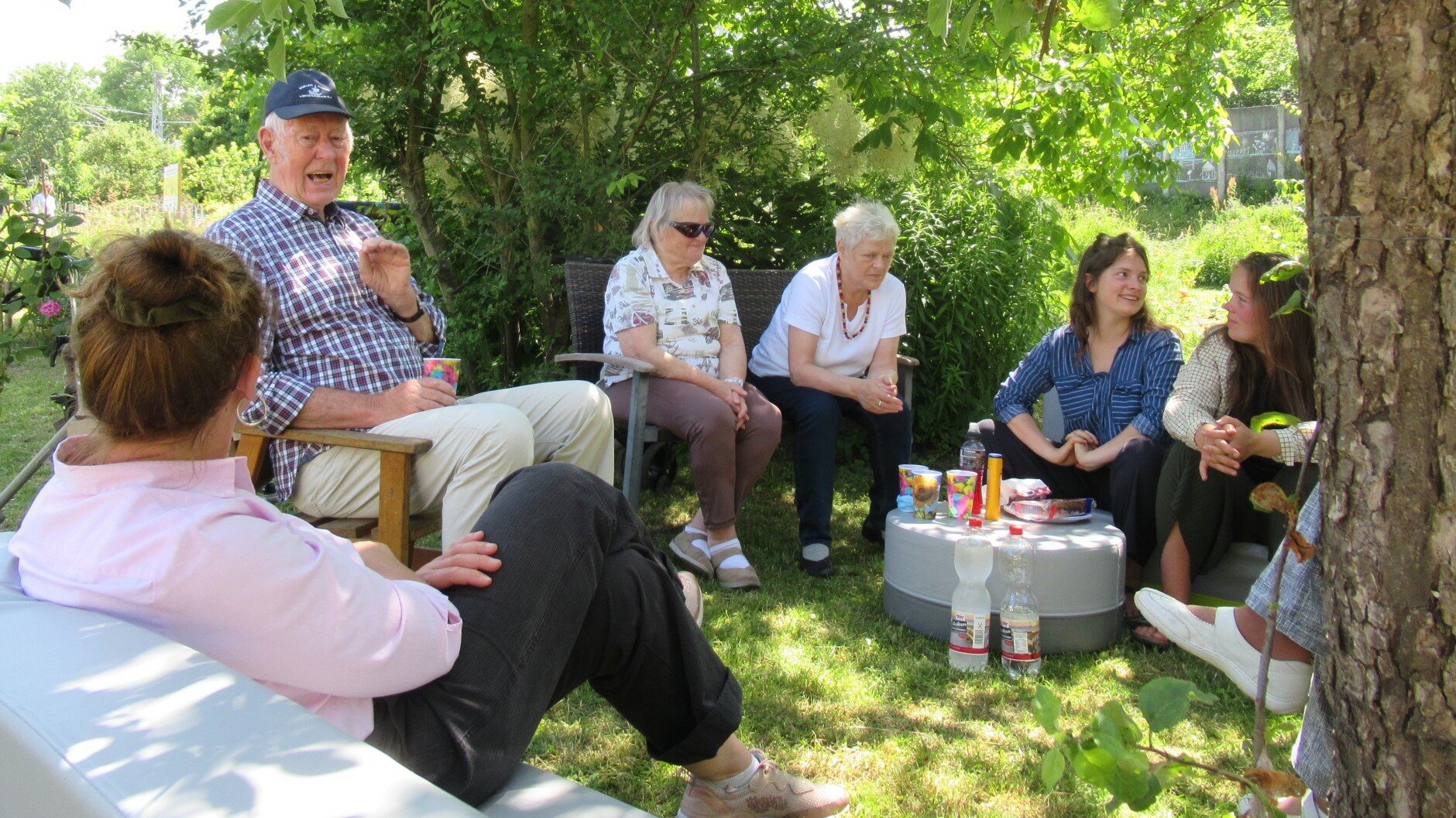 Sechs Gärtnerinnen und Gärtner sitzen in einem Halbkreis im Garten, unterhalten sich und genießen eine Trink- und Vesperpause.