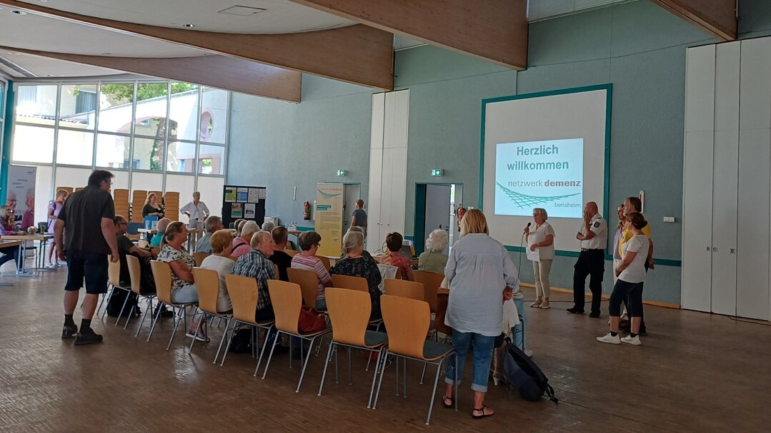 Foto: Menschen bei einer Veranstaltung in einem Saal mit Blick auf eine großen Leinwand, vor der mehrere Personen stehen, eine mit Mikrofon.
