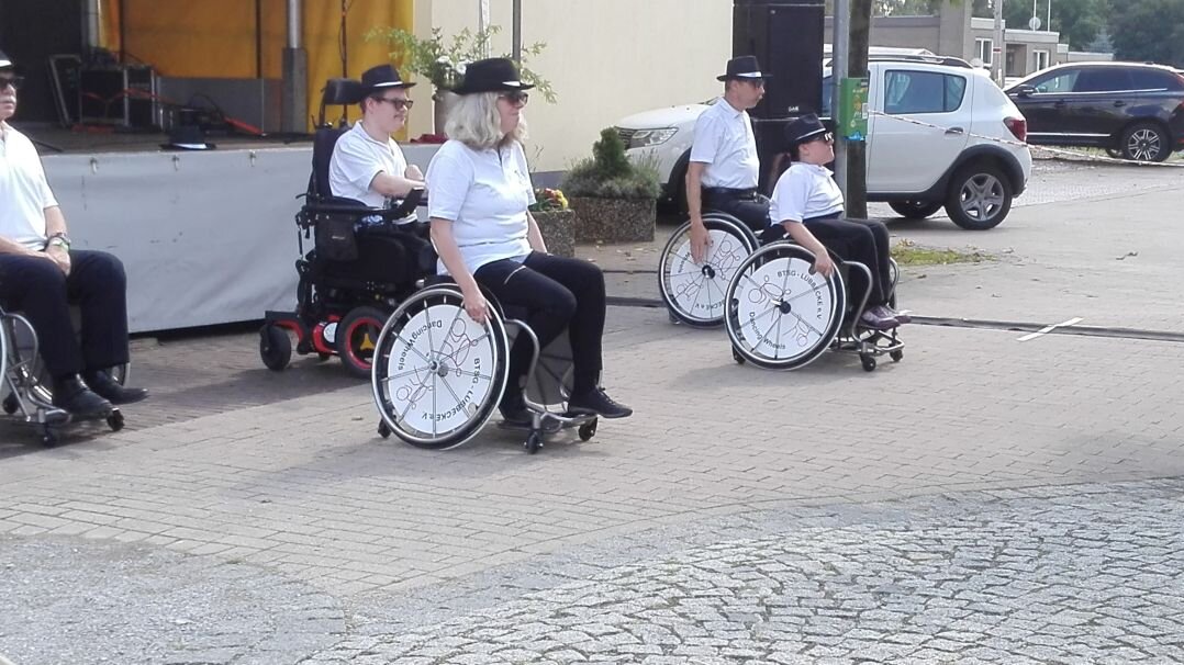 Auf diesem Foto sieht man vier Menschen unterschiedlichen Alters, die einen Tanz aufführen. Alle sitzen im Rollstuhl. 