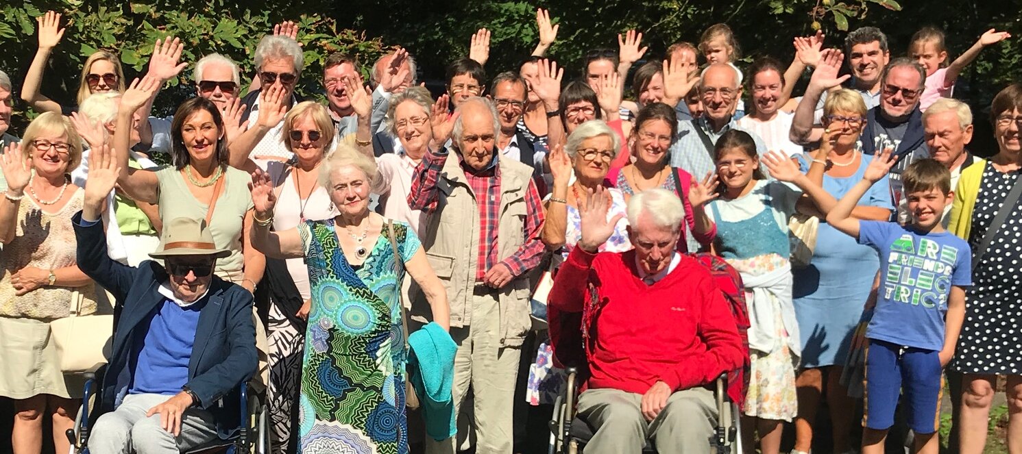 Gruppenbild von Menschen unterschiedlichen Alters, die in die Kamera winken