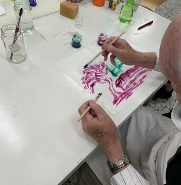 An einem Tisch malt eine ältere Person mit Aquarellfarben