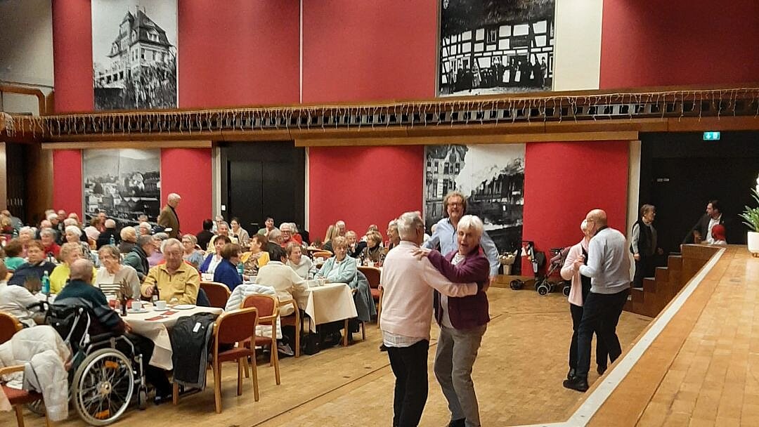 Foto: Senior*innen sitzen an langen Tischen in einem Saal mit roten Wänden. Vor der Bühne sieht man zwei Tanzpaare im Seniorenalter.