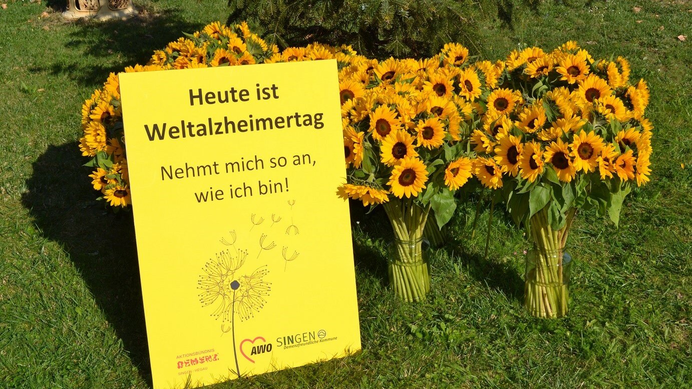 Vasen mit Sonnenblumen. Davor steht ein gelbes Plakat mit der Aufschrift "Heute ist Weltalzheimertag!"