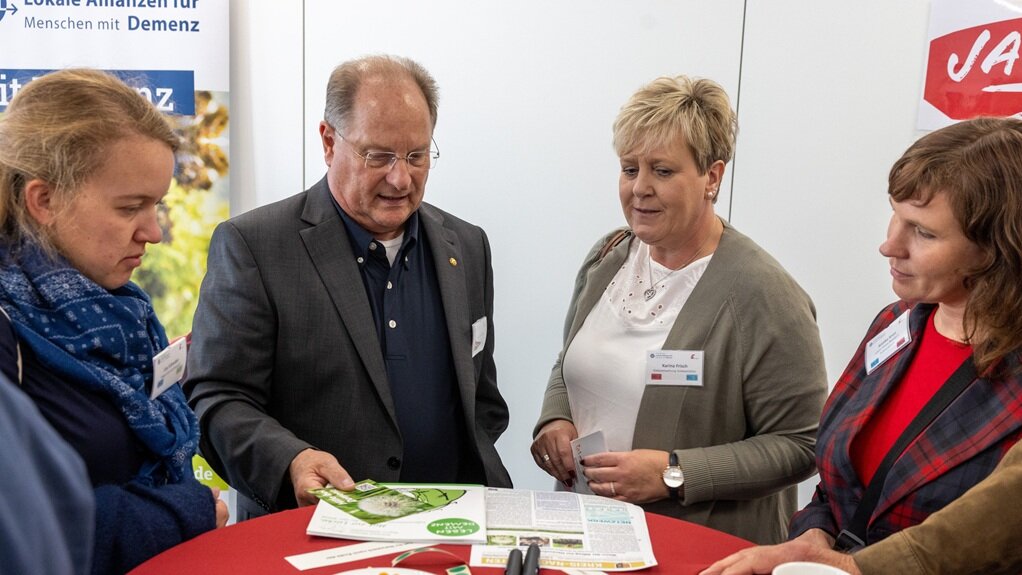 Informeller Austausch der Lokalen Allianzen beim Get-together  Foto: Vier Personen betrachten einen Flyer am Stehtisch.