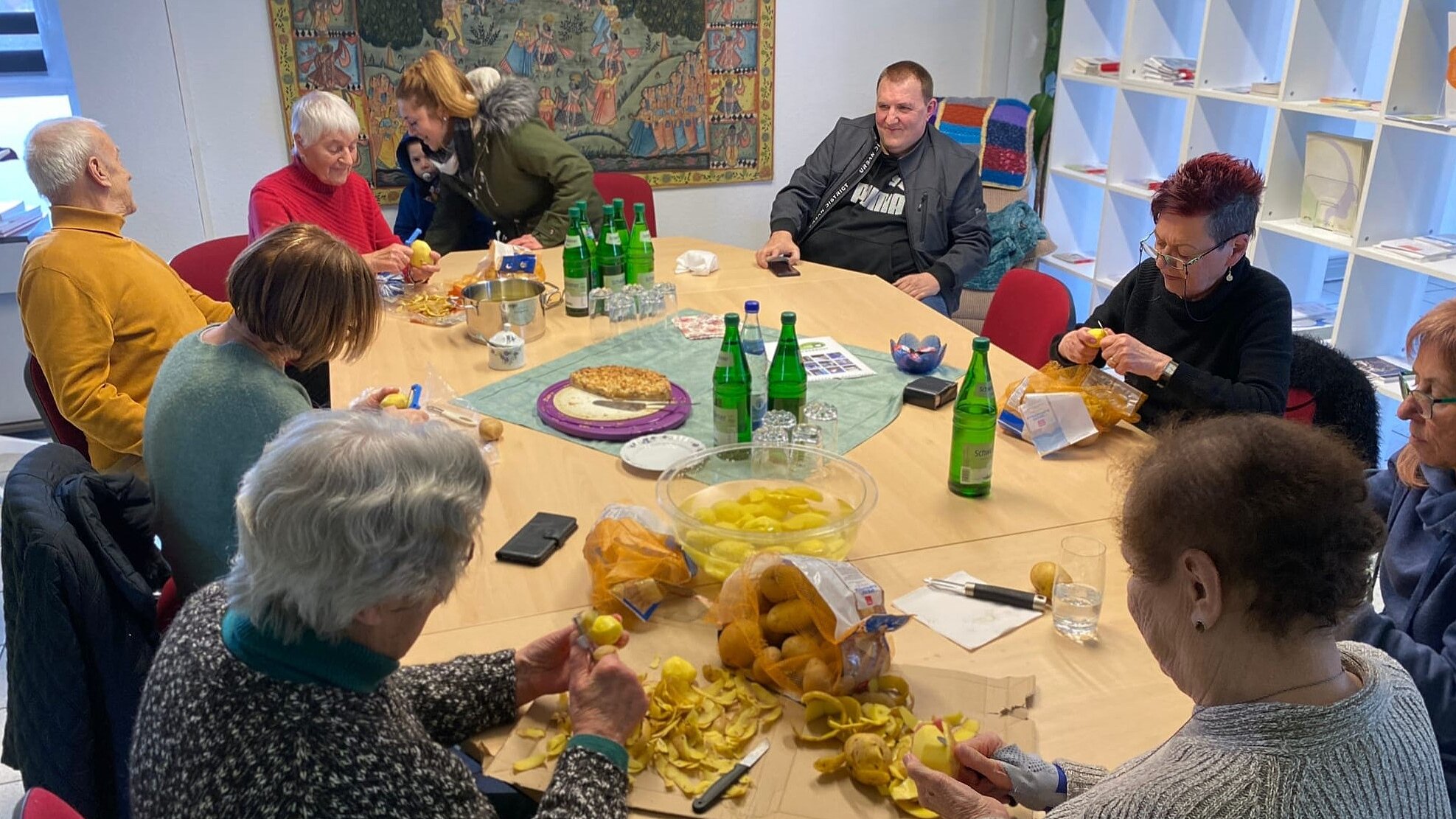 Gemeinsam kochen – gemeinsam wirken: Jung und Alt bereiten gemeinsam eine warme Mahlzeit für den „Kältebus“ zu. Kinder, Jugendliche, Menschen mit Demenz und Senior*innen erleben dabei gelebte Teilhabe, Zusammenhalt und Solidarität. Foto: 10 Menschen aus verschiedenen Generationen sitzen an einem großen Tisch und schälen Kartoffeln