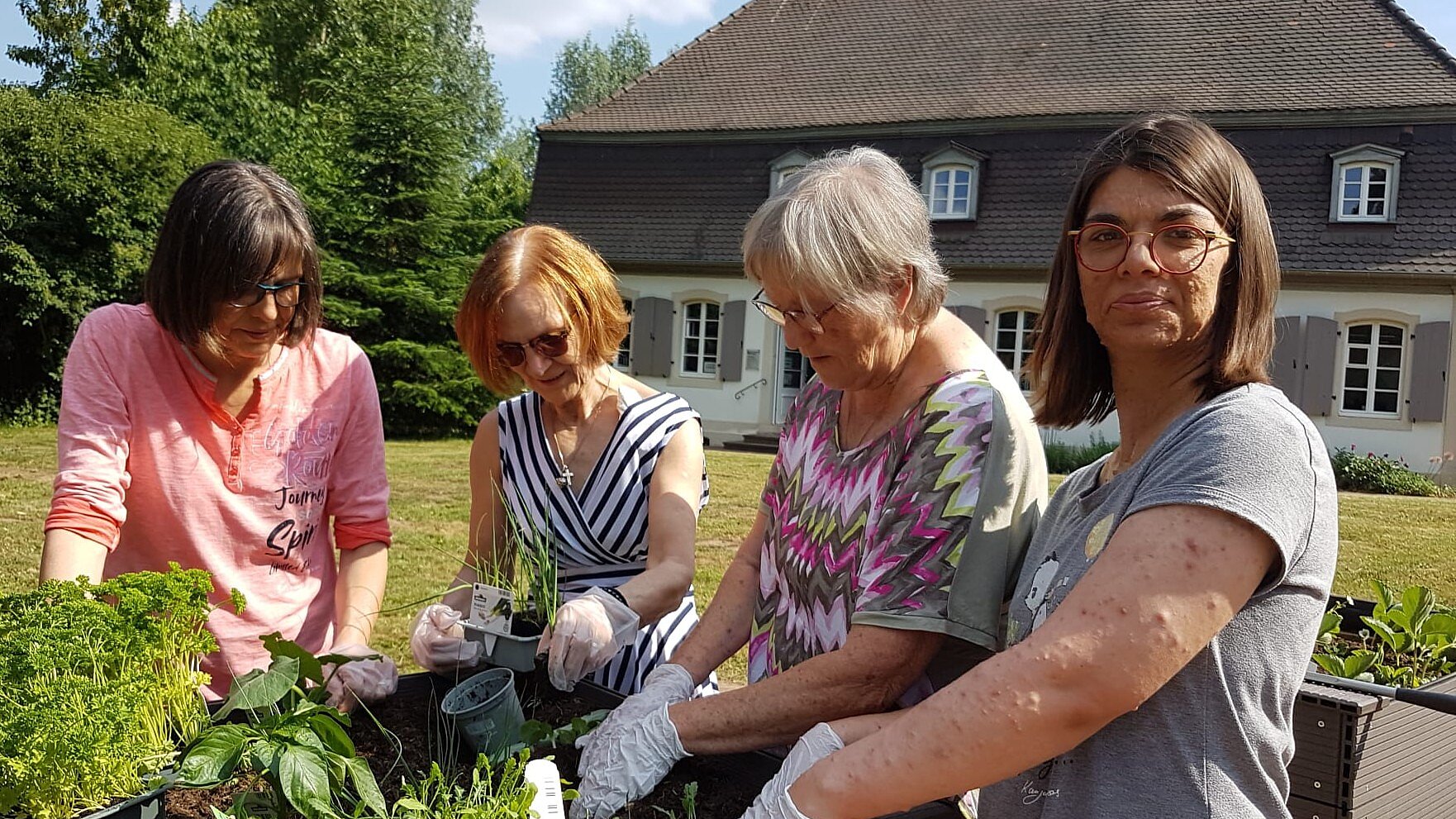 Foto: 4 Frauen aus unterschiedlichen Generationen gärtnern an einem Hochbeet vor einem alten Haus.