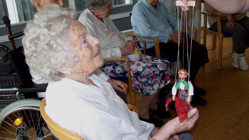 Wenn Seniorinnen und Senioren nicht mehr ins Theater gehen können, kommt das Theater zu ihnen: das mobile Marionetten-„Theater aus der Truhe“ zu Gast im Stiftungsdorf Hollergrund Foto: Eine sitzende ältere Dame mit einer Marionette.