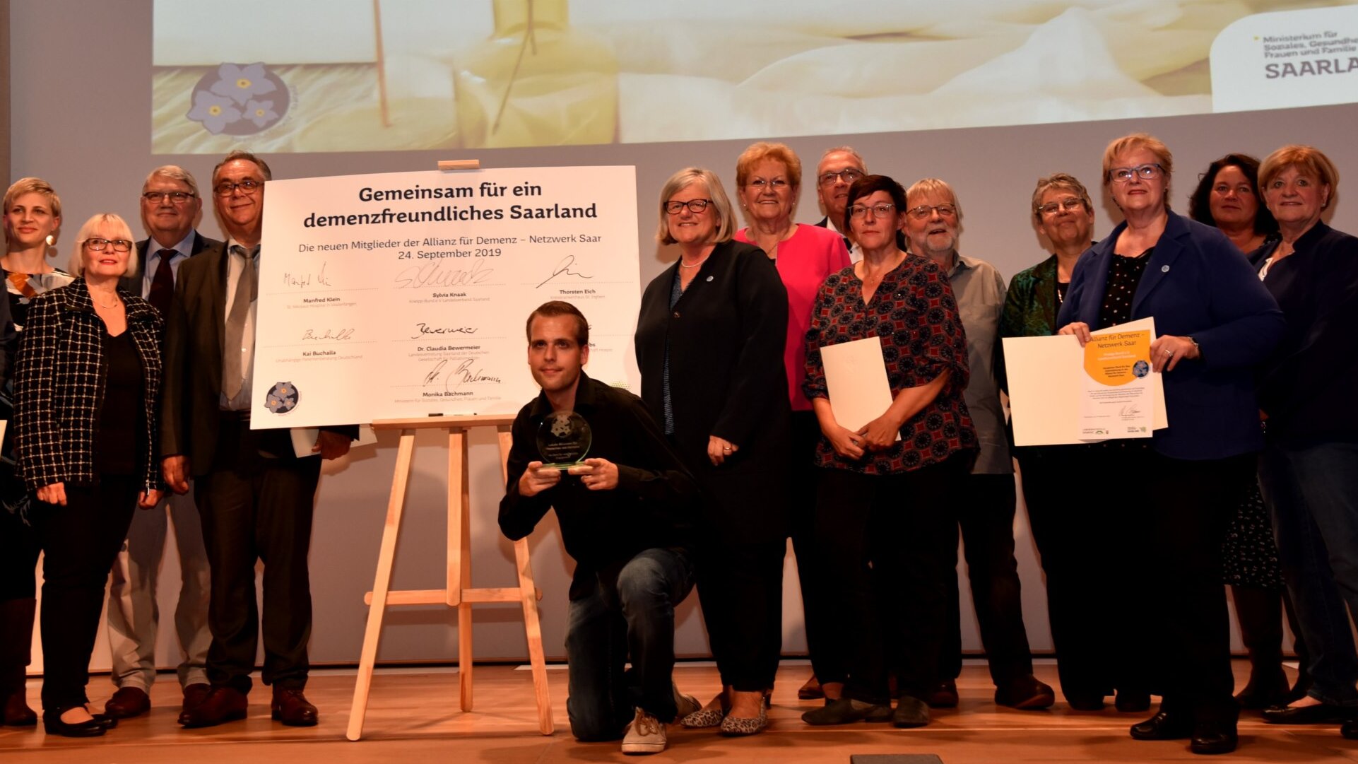 Auszeichnung des Projekts durch das Sozialministerium in Saarlouis, 2019 Foto: Die Projektbeteiligten des Vereins zur Förderung der Dorfentwicklung Hoof e.V. präsentieren auf einer Bühne ihre Auszeichnung