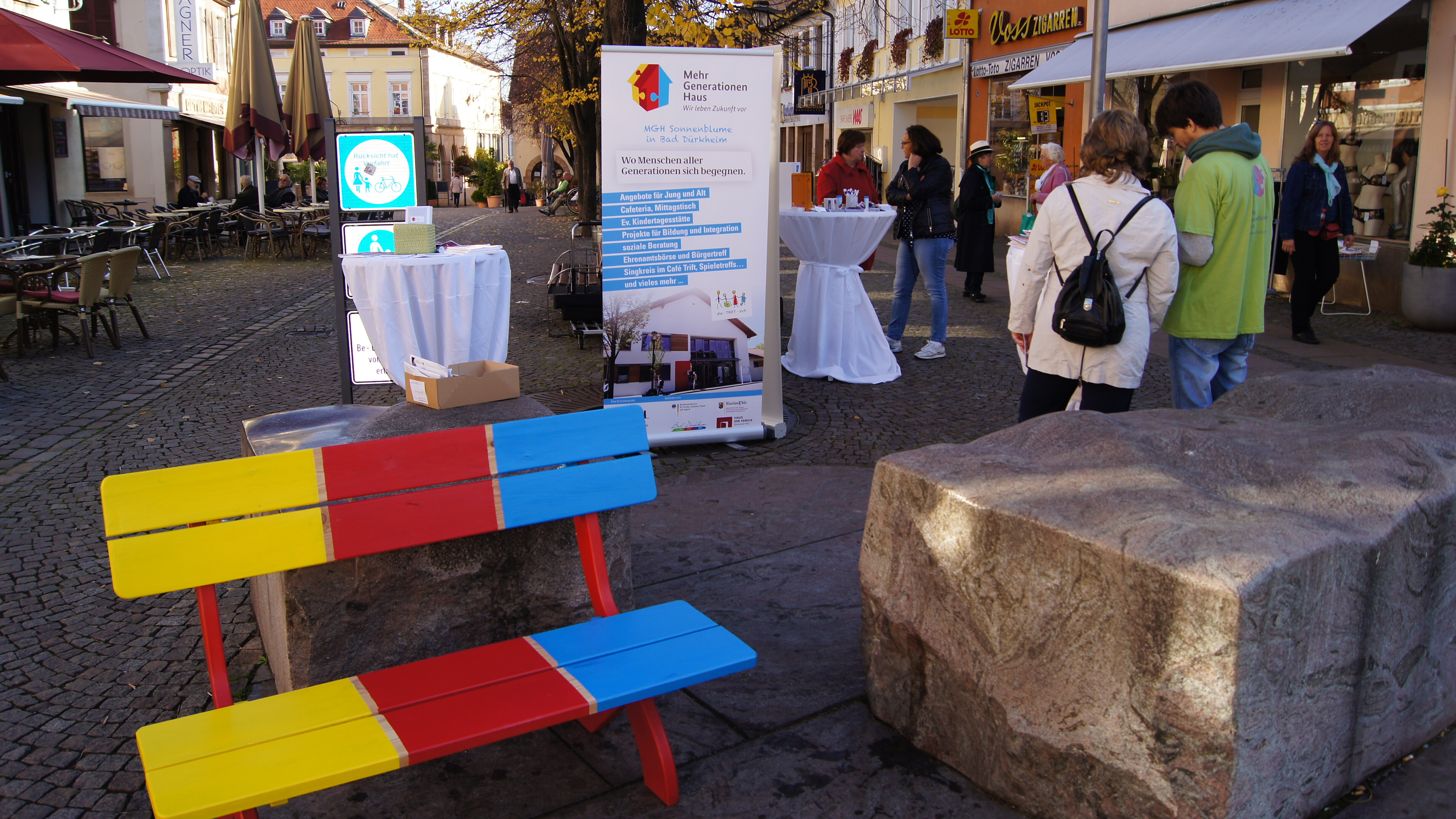 Foto: Eine draußen stehende bunte Sitzbank. Daneben ist ein größerer Steinblock. Im Hintergrund sind Personen, Stehtische und ein Roll Up vom Mehrgenerationenhaus.